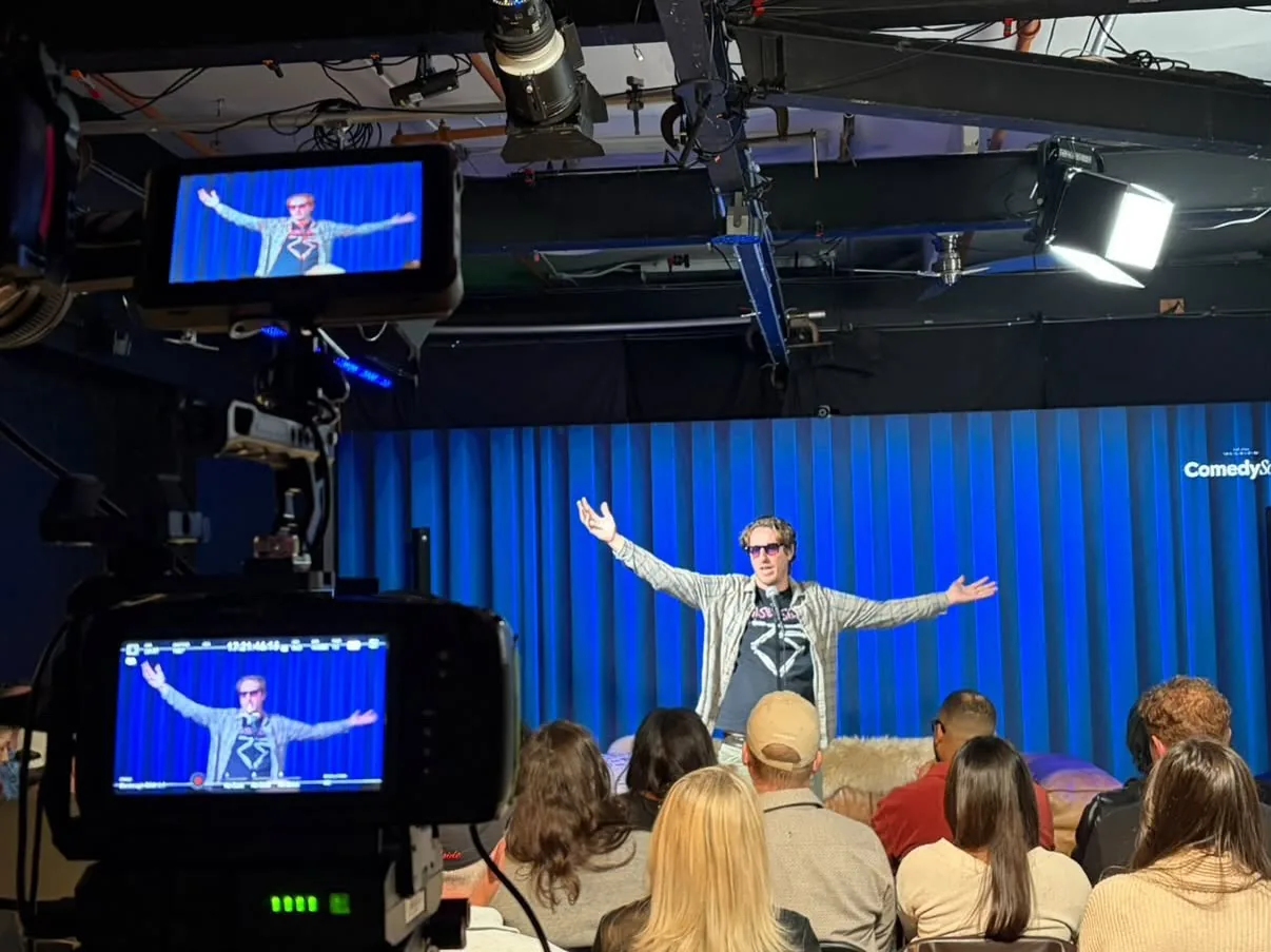 A comedian on stage at Comedy Social, the crowd in front and the camera monitor rolling